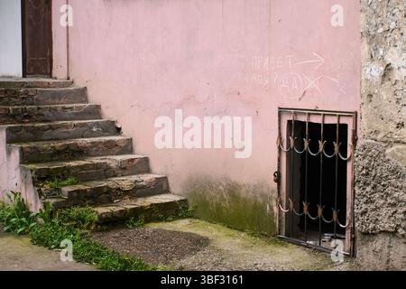 Un frammento di un vecchio edificio con una scalinata in pietra che conduce verso l'alto e una finestra barrata sotto. Pareti squallide, vegetazione Foto Stock