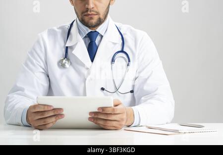 Primo piano delle mani del medico maschile usando un computer tablet. Medico in uniforme medica bianca con stetoscopio seduta alla scrivania. Tecnologia moderna in examina Foto Stock