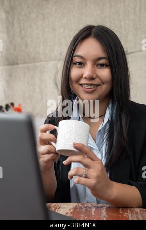 Una giovane donna sicura di sé ama il caffè mentre lavora in remoto, sorridendo naturalmente al suo notebook in uno spazio di lavoro moderno. Luminoso, caldo e conveniente. Foto Stock