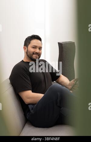 Un uomo ben vestito in un abbigliamento completamente nero, che include un maglione e pantaloni, si siede comodamente in una moderna sala ufficio. Concentrato sul suo tablet, l'app Foto Stock
