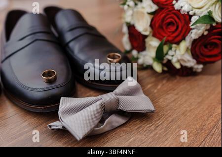 Una scena nuziale ben organizzata presenta eleganti scarpe nere, anelli dorati a simboleggiare l'impegno, un papillon grigio testurizzato e un bouquet straordinario Foto Stock