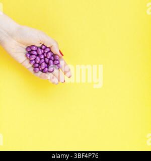Donna mano azienda viola pillole manciata isolato in grassetto su sfondo giallo con copia spazio. Modello per la bellezza femminile blog social media. Hea femmina Foto Stock
