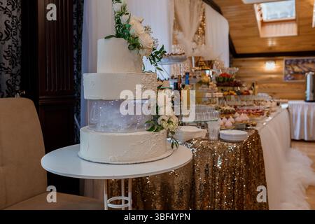 Una bellissima torta nuziale bianca a quattro piani decorata con delicati fiori bianchi è il fulcro di una splendida scena di ricevimento, caratterizzata da un sequ dorato Foto Stock