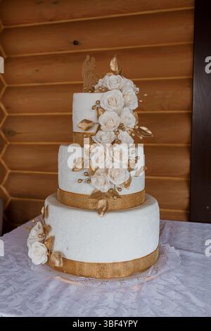 Una stupenda torta nuziale bianca a tre piani decorata con nastro d'oro e delicate rose si erge splendidamente sullo sfondo di una rustica parete di tronchi Foto Stock