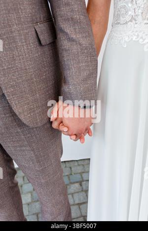 Uno scatto ravvicinato mostra una coppia di sposi che si tiene per mano durante la cerimonia di nozze, con lo sposo che indossa un abito marrone in tweed e la sposa in bianco Foto Stock