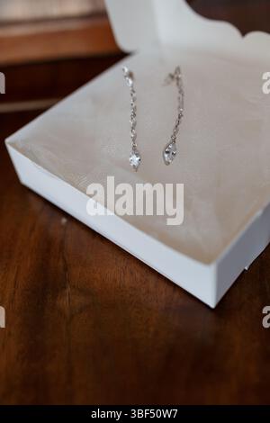 Un paio di scintillanti orecchini pendenti in argento con scintillanti pietre di zirconia cubica si trova all'interno di un immacolato portagioie bianco posizionato su un ricco legno scuro Foto Stock