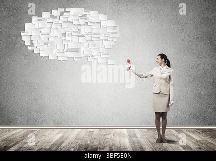 Giovane donna che tiene in mano il telefono rosso e bolle di voce su sfondo grigio. Operatore di call center in tuta da lavoro bianca. Conversazione di lavoro e inter Foto Stock
