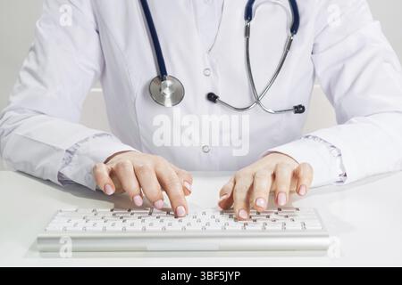 Primo piano medico che digita sulla tastiera del computer. Medico donna in camice bianco con stetoscopio seduto alla scrivania. Diagnosi e trattamento medico professionale Foto Stock