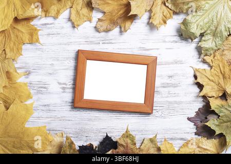 La cornice rettangolare vuota si trova su una scrivania in legno vintage con un brillante fogliame autunnale. Stendere in piano con foglie autunnali su una superficie di legno bianca. Fronte semplice Foto Stock