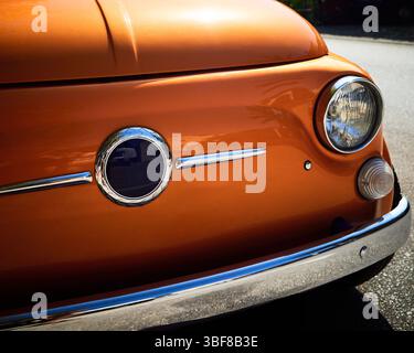 Primo piano di un'auto d'epoca arancione con paraurti cromato e faro rotondo, parcheggiata alla luce del sole su una strada mediterranea. Foto Stock