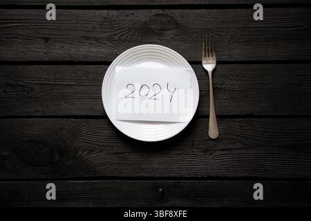 L'iscrizione 2024 su un pezzo di carta in un piatto con una forchetta su una superficie in legno nero, piano aziendale, obiettivi, tempi e scadenze Foto Stock