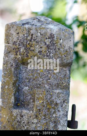 Primo piano di palo in pietra con inserto e cerniera in metallo; pietra grigio chiaro con lichene verde e nero, sfondo fogliato sfocato. Foto Stock