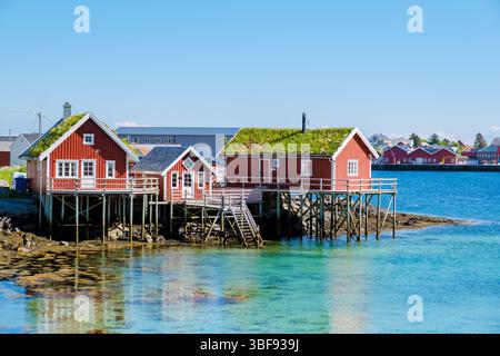 Le colorate capanne rosse arroccate su palafitte si riflettono nelle limpide acque della costa norvegese di Reine Lofoten sotto un cielo blu brillante. Foto Stock