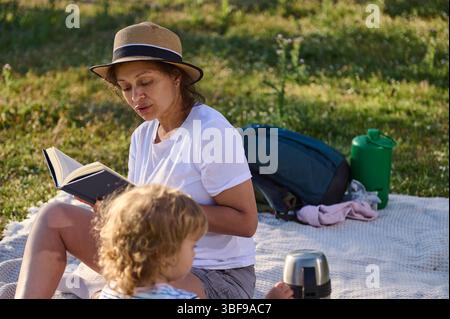 Una madre ama trascorrere del tempo all'aperto leggendo un libro al proprio bambino in una calda giornata di sole, mettendo in risalto il legame familiare e abbracciando la serenità della natura. Foto Stock