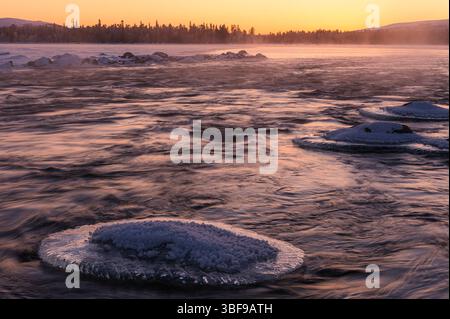 Uno splendido tramonto invernale dipinge il cielo in vivaci sfumature sopra un fiume ghiacciato in Norvegia. Le formazioni di ghiaccio galleggiano sull'acqua, creando un'atmosfera tranquilla Foto Stock