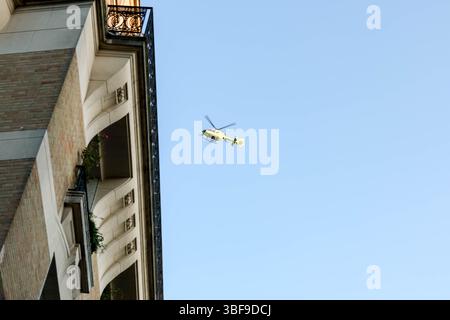 Un elicottero giallo brillante viene catturato a metà volo contro un vasto cielo azzurro, con le punte dei rami d'albero visibili in primo piano, i nastri trasportatori Foto Stock