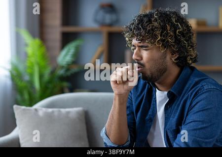 Un uomo dai capelli afro, che tiene il pugno alla bocca, tossisce seduto su un divano. Sembra che possa avere un raffreddore o una malattia simile. Foto Stock