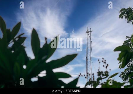 Una suggestiva torre di comunicazione si erge su un cielo blu luminoso pieno di nuvole wispy, circondata da una vivace vegetazione verde che mostra tecnol rurale Foto Stock