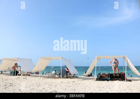 Spiaggia con tende bianche a baldacchino e bagnanti che si godono una giornata di sole in riva al mare. Cielo azzurro e acque turchesi in un ambiente costiero tropicale. Foto Stock