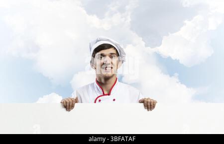 Giovane chef maschile con cartello bianco. Splendido chef in uniforme bianca e rossa su sfondo blu. Annuncio delle lezioni di cucina. Cateri professionali Foto Stock