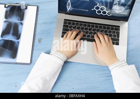 Vista dall'alto delle mani del medico che scrivono sul computer portatile. Terapista in camice bianco seduto alla scrivania di legno blu. Esame e consultazione in clinica. Medico Foto Stock