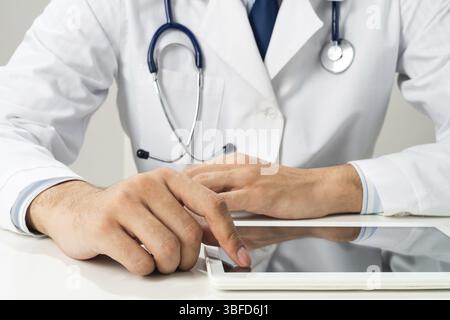 Primo piano delle mani del medico maschile usando un computer tablet. Terapeuta in abito medico bianco con stetoscopio seduto alla scrivania. Tecnologia moderna nella diagnosi Foto Stock