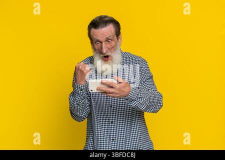 Vecchio anziano che usa lo smartphone per celebrare il successo della vittoria vittoria gioca all'applicazione del casinò della lotteria, simulatore di guida di corsa sparatutto videogiochi. Anziano nonno barbuto isolato su sfondo giallo Foto Stock