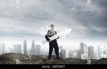 Giovane imprenditore di successo nel soddisfare mantenendo la grande freccia bianca e rivolto lontano mentre in piedi tra le rovine con vista città e cielo nuvoloso sul backgr Foto Stock