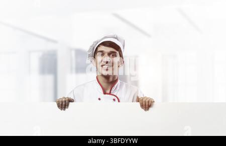 Giovane chef maschile con cartello bianco bianco bianco. Splendido chef in uniforme bianca e rossa su uno sfondo sfocato della stanza. Annuncio delle lezioni di cucina. Pro Foto Stock