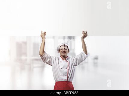 Giovane chef maschile con un cartello vuoto sopra la testa. Chef in uniforme bianca e rossa su sfondo sfocato in camera chiaro. Annuncio delle lezioni di cucina. Confessare Foto Stock