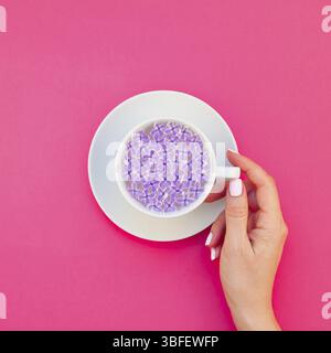 Quadrato piatto concetto laici top view tazza bianca bellissimi fiori lilla galleggianti in acqua infusa e donna mano millenaria sfondo rosa spazio copia mini Foto Stock