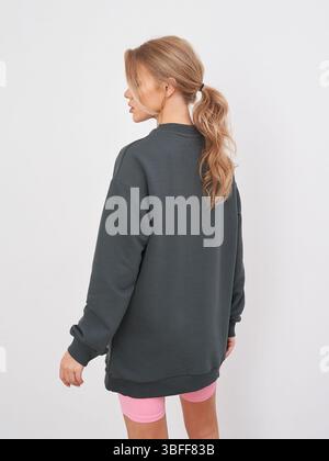 Donna con capelli lunghi in coda di cavallo, rivolta verso l'esterno, che indossa felpa scura e pantaloncini rosa. Abbigliamento casual, luce soffusa su di lei, espressione neutra Foto Stock