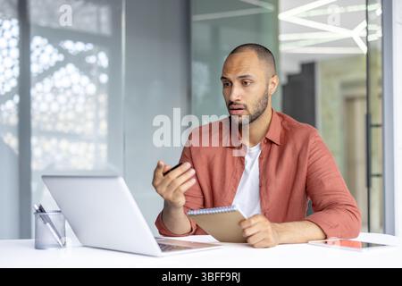 Un uomo concentrato conduce una videochiamata nel suo ufficio, tenendo in mano un blocco note, interagendo attivamente con qualcuno dall'altra parte. Foto Stock