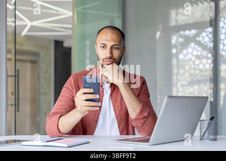 Un uomo serio tiene uno smartphone a una scrivania con un laptop, un notebook e un portapenne, possibilmente funzionante. Foto Stock