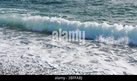 Infrangere le onde su una spiaggia rocciosa, formando spruzzi e spruzzi. L'oceano è calmo e le onde sono piccole. L'acqua è una bella sfumatura di blu Foto Stock