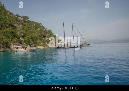 Una tranquilla baia in Turchia ospita numerosi goleti tradizionali all'ancora, con i loro alti alberi che creano una silhouette suggestiva contro il verde lussureggiante rugge Foto Stock