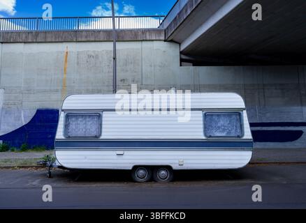 Carovana abbandonata con pneumatici sgonfi parcheggiata sotto un ponte di Berlino, evocando solitudine urbana e contrasto. Foto Stock