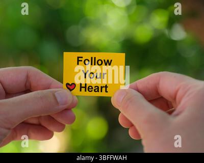 Una donna che tiene in mano un biglietto giallo con la frase Segui il tuo cuore scritto sulle sue mani su uno sfondo fogliame. Primo piano. Foto Stock