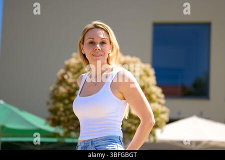Donna bionda sicura con canotta bianca e jeans in posa all'aperto alla luce del sole. Foto Stock