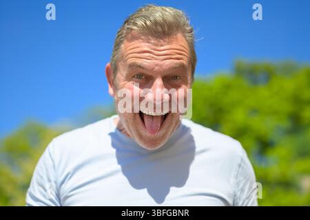 Un uomo anziano allegro e giocoso tira fuori la lingua sorridendo in grande, godendosi una giornata di sole all'aperto. Foto Stock