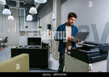 Giovane uomo che utilizza un computer portatile mentre si trova accanto alla stampante in un ufficio moderno durante il giorno Foto Stock
