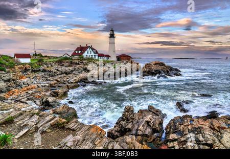 Portland Head Light si erge in modo prominente sulla costa rocciosa di Cape Elizabeth, Maine, Stati Uniti, incorniciata da spettacolari onde oceaniche e colorati cieli serali. Lo storico faro e i suoi edifici dal tetto rosso contrastano splendidamente con la costa di granito naturale e il mare aperto oltre Foto Stock