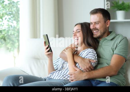 Coppia felice che si diverte a guardare i contenuti multimediali sul telefono cellulare seduto sul divano di casa Foto Stock