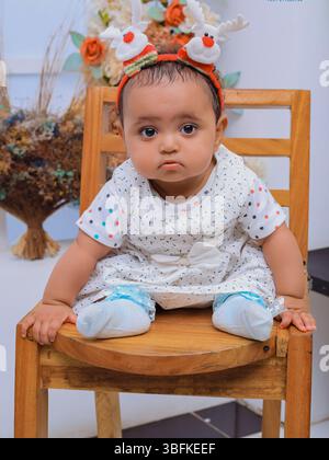 Un simpatico bambino è seduto su una panchina bianca, vestito con un vestito colorato composto da una t-shirt azzurra con colletto rosa simile a petali e rosa Foto Stock