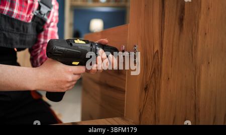 Assemblaggio di mobili e fissaggio di accessori, installazione di una cerniera per mobili con un trapano avvitatore. primo piano Foto Stock