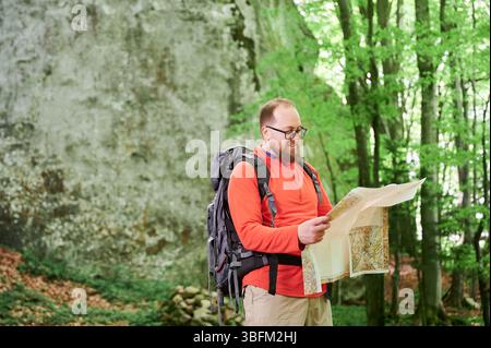 Un turista sorridente e barbuto studia la mappa mentre si trova di fronte a una grande roccia nella fitta foresta. Viaggiatore felice con zaino e occhiali grigi, pianificazione di avventure all'aperto, escursioni o arrampicate. Foto Stock