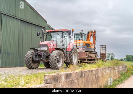 Vista anteriore del trattore agricolo con rimorchio e scavatore sul lato posteriore del rimorchio. Il trattore è parcheggiato davanti a un grande corr verde Foto Stock