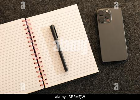 Composizione piatta e minimalista con un notebook a spirale aperto, una penna nera e uno smartphone moderno su un tappetino da scrivania grigio scuro. Ideale Foto Stock