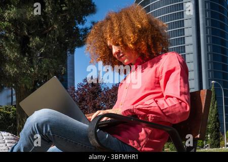 Un giovane sicuro di sé con i capelli rossi ricci siede su una panchina che lavora sul suo laptop in un ambiente urbano. I moderni edifici per uffici dietro di lui mettono in risalto il di Foto Stock