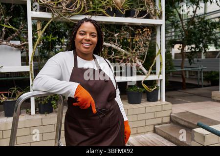 Una donna afroamericana con trecce irradia gioia mentre si occupa delle piante in una serra. Foto Stock
